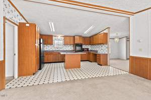 Spacious kitchen with brown wooden cabinets, black appliances, and a central island. The room has a white tiled floor with black accents and floral wallpaper. An adjoining room is visible with beige carpeting and a light fixture.