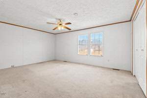 An empty room with beige carpet and white walls. A ceiling fan with a light fixture is centrally placed. Two large windows are on the right wall, allowing natural light. A closed white door is to the right.