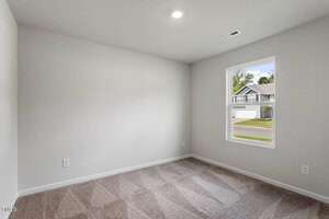 A small empty room with light gray walls and beige carpet. The ceiling has one recessed light. A window on the right wall reveals a street view with houses.