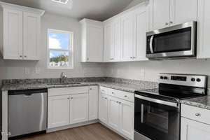 A modern kitchen with white cabinets and granite countertops. It features stainless steel appliances, including a microwave, stove, and dishwasher. A small window above the sink allows natural light to enter. The floor is made of wood.