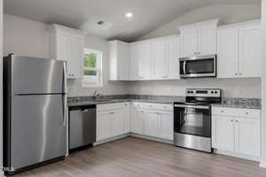 Modern kitchen with white cabinets, stainless steel appliances, and dark granite countertops. Includes a refrigerator, dishwasher, and oven with a microwave above. Light wood flooring and a small window near the sink.