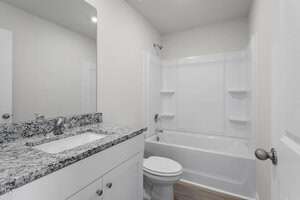 A bathroom with a white bathtub and shower, granite countertop sink, and a toilet. The walls are light-colored, and the floor is wood-patterned. A mirror is above the sink, reflecting the light and ceiling.