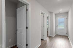 A hallway with light wood flooring and light gray walls. Several open doors lead to different rooms, one showing a glimpse of a carpeted area. At the end of the hallway, there is a white door with a frosted glass panel.
