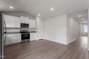 A modern kitchen with white cabinets, stainless steel appliances including a microwave and oven, and light wood flooring. The space opens into a hallway leading to an adjacent room. Recessed lighting is installed in the ceiling.