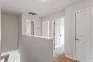 Hallway with beige walls and carpeted floor. There is a white door on the right, an open door leading to another room, and an additional doorway on the left. A half-wall is shown in the foreground. Ceiling lights illuminate the space.