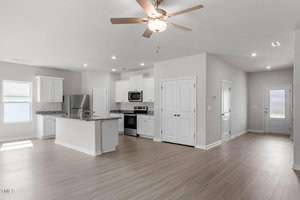 Open-concept kitchen and living area with light wood flooring. The kitchen features white cabinets, a central island with granite countertops, stainless steel appliances, and a ceiling fan. A door leads to an adjacent hallway.