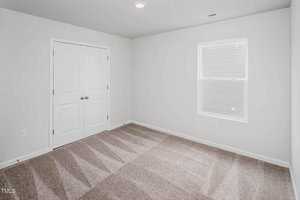 Empty room with white walls and a beige carpet. A closed white double door is on the left, and a single window is on the right wall. The room is well-lit with a ceiling light. The floor shows visible vacuum lines.