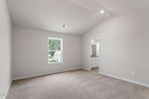 Empty room with beige carpet, white walls, and a sloped ceiling. A single window shows a view of trees and a neighboring house. An open doorway leads to another room with visible white cabinets. Recessed lighting is installed in the ceiling.