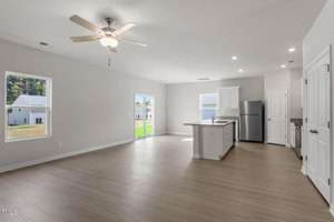A spacious, unfurnished open-plan living area and kitchen with light wood flooring. The kitchen features white cabinets, a central island, and a stainless steel refrigerator. Large windows and a sliding glass door provide natural light.