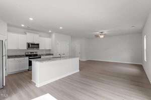 Spacious, empty kitchen and living area with white walls and light wood floors. The kitchen features white cabinets, a central island with granite countertop, stainless steel appliances, and recessed lighting. The living area has a ceiling fan and a door.