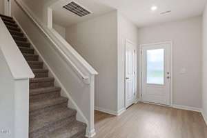A small foyer area with light wood flooring. A carpeted staircase is on the left, leading upward. A white door with frosted glass is straight ahead, and a closed white interior door is on the right. The walls are painted light gray.