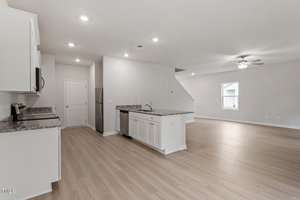A modern, open-concept interior with a kitchen featuring white cabinets, granite countertops, and stainless steel appliances. The space flows into a large, light wood-floored living area with a ceiling fan. A staircase is visible in the background.
