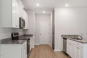 A modern kitchen with white cabinets, stainless steel appliances, and granite countertops. There is a sink with a faucet on the right and a refrigerator next to a closed white door at the back. The floor is light wood.