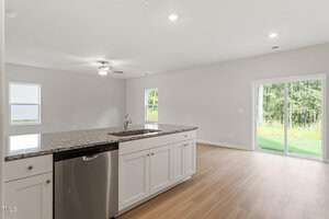 Modern kitchen with a large island featuring a granite countertop, white cabinets, and a stainless steel dishwasher. The open floor plan leads to a living area with a ceiling fan and glass sliding doors opening to a grassy outdoor space.