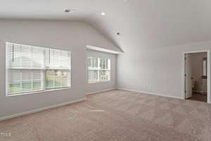 A bright, empty room with light beige carpet and white walls. There are two large windows with blinds on the left side, a vaulted ceiling, and an open doorway leading to a bathroom on the right.