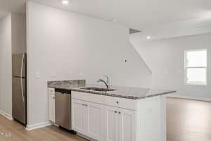 A modern kitchen features white cabinets, granite countertops, and a stainless steel dishwasher. A refrigerator is positioned against the wall. The area opens into a spacious, well-lit living room with light wood flooring.