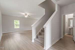 An empty room with light wood flooring and a beige carpeted staircase leading upward. A ceiling fan is mounted on the light-colored ceiling. To the right, there is a door leading to a small bathroom with a sink and mirror. A window is visible on the far wall.