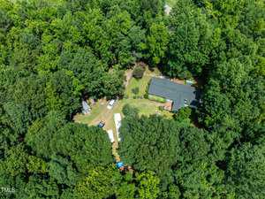 An aerial view of a house surrounded by dense green trees. The property includes a manicured lawn, a long driveway, and several structures, possibly storage or recreational. The surrounding forested area is lush and verdant.