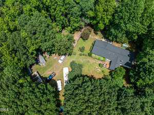 Aerial view of a house surrounded by dense green trees. The property includes a black-roofed building, vehicles, and assorted outdoor items. A grass-covered yard is visible, along with some shrubs and pathways.