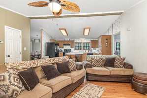 Living room with two brown sofas, patterned cushions, and a ceiling fan with leaf-shaped blades. An open view to the kitchen shows a black fridge, wooden cabinets, and a breakfast bar with stools. Walls have floral wallpaper. Wood flooring is visible.