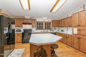 A kitchen with wooden cabinets and a central island with a decorative horse carving. Black appliances are present, including a refrigerator and stove. The walls have a floral wallpaper, and the ceiling has fluorescent lighting.