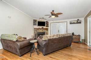 A living room features two brown sofas, a stone fireplace with a mounted TV above, and wooden floors. A ceiling fan with leaf-shaped blades hangs overhead. French doors are on the far wall, and a wooden side table is on the right.