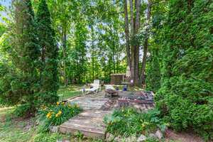 A wooden deck in a lush backyard contains two white lounge chairs and a fire pit. Tall evergreens and other plants surround the area. In the background, there is a closed umbrella and a small rustic structure.