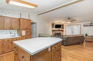 A kitchen with wooden cabinets and a white countertop island. The adjacent living room has a brown sofa, a stone fireplace, and a mounted TV. Wooden flooring extends throughout, with a ceiling fan and double doors in the background.