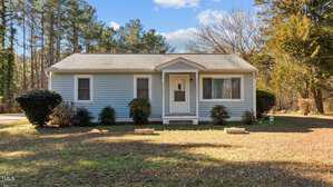 A small, single-story house on Henderson's tranquil 1625 Gun Club Road features light blue siding and a gray roof. The front showcases two windows and a central door with a modest overhang. Bushes flank the entrance, while trees create a serene backdrop.