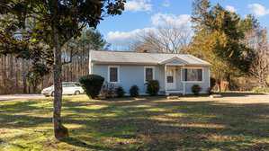 Small single-story house with light blue siding and a gray roof, set on a grassy lawn with trees and shrubs surrounding it. A white car is parked on a paved driveway to the left. The background features tall, leafless trees under a partly cloudy sky.