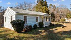 A small, single-story house with light blue siding and a gable roof. The front porch has a white door and two windows. Bushes are planted in the yard, and there are trees in the background under a clear sky.
