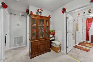 A hallway with a wooden cabinet containing glass doors displaying floral items. Red flowers adorn the walls and doors. The hallway leads to a bathroom with towels and decor. A cardboard box with folded blankets sits on the floor.