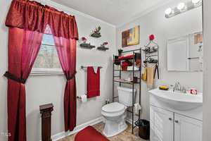 A small bathroom with a white sink and toilet. Red towels, curtain, and rug provide accents. Shelves hold decor, including red flowers and candles. A window with a red curtain is on the left. Walls are painted light gray, and a small artwork is hung.