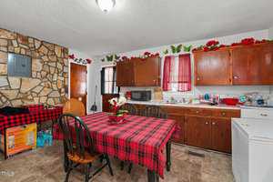 A kitchen with wooden cabinets, a stone accent wall, and red plaid tablecloth on a dining table with four chairs. Red decorations and butterfly accents line the cabinets. A microwave, dishwasher, and cleaning supplies are visible.