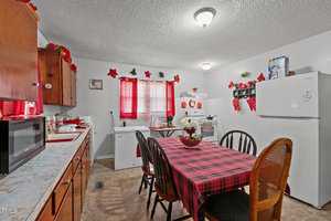 Kitchen with a plaid tablecloth on a rectangular table surrounded by chairs. Wooden cabinets line the wall to the left, a refrigerator to the right. A window with red curtains, stove, and small decor items are visible. Ceiling light illuminates the room.