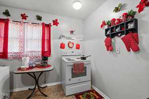 A small kitchen with white walls and a red theme. There's a stove with red pot holders, a wall shelf with red mugs, and red poinsettia decorations. A table holds a coffee maker, snacks, and paper towels, under a window with red curtains.