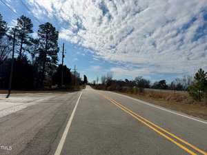 A rural two-lane road with yellow lines runs straight ahead under a partly cloudy sky. Trees line the road on both sides, and power lines are visible. The scene is peaceful and open, with light traffic and natural surroundings.