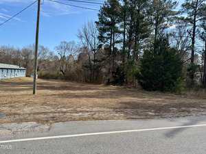 A rural scene featuring a paved road running alongside a small grassy area. To the left, there's a long, low building with a blue exterior. On the right, a cluster of tall trees borders the property. The sky is clear and blue.