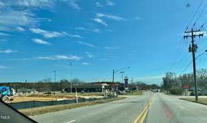 View of an empty road with a construction site on the left, featuring a blue excavator. In the background, there is a restaurant and fast-food establishment with signage. The sky is clear with scattered clouds.