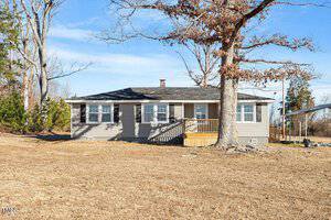 A small, single-story house at 2041 Grassy Creek Virgilina Road features a gray exterior and a covered front porch. It sits in a grassy area near Oxford, with a large tree in the foreground. The sky is clear and blue, and the yard is surrounded by sparse trees.