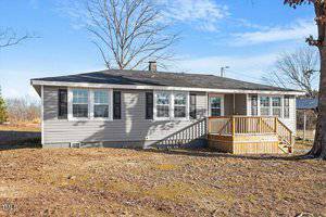 A single-story house with light gray siding and a dark roof. It features a small wooden porch with stairs leading up to the front door. Bare trees and a clear blue sky are in the background. The ground is covered with dry grass and patches of dirt.