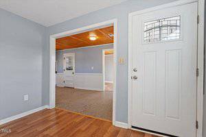 The image shows an interior entryway with light wood flooring. A white door with a decorative glass inset is on the right. The entry leads to a carpeted area with wood-paneled ceiling, separated by an open wall. Walls are painted light blue.