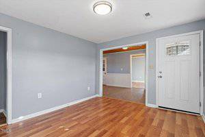 A room with light blue walls and wooden flooring. A white door with a glass panel is on the right. A doorway without a door leads to another room with similar flooring. The ceiling light is turned on. A vent is visible near the ceiling.