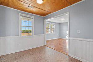 A room with light gray walls and white wainscoting features a tiled floor and a wooden ceiling. It opens into another room with hardwood flooring, large windows, and similar walls. Both rooms have ceiling lights and ample natural light.