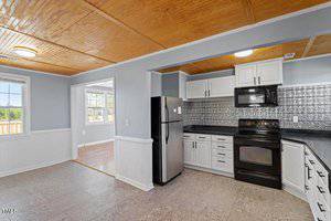 A kitchen with light gray walls, featuring white cabinets, a black refrigerator, stove, and microwave. The backsplash is decorative metal, and the ceiling has wooden paneling. A window provides natural light, and a doorway leads to an adjoining room.