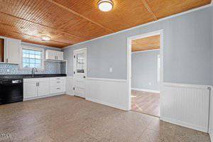 A kitchen with light blue walls, white cabinets, and a wooden ceiling. There is a black dishwasher and a black countertop with a stainless steel sink. A door leads to an adjoining room with wood flooring.
