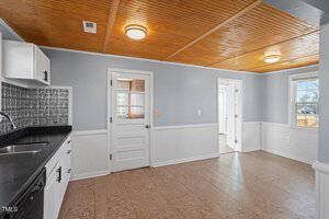 A kitchen with a wooden ceiling and tiled floor. It has white cabinets, a black countertop, and a silver backsplash. There is a sink, a white door, and two windows allowing natural light. The walls are painted light blue with white wainscoting.