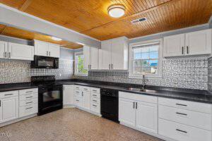 A modern kitchen with white cabinets and a black countertop. The backsplash features a decorative silver tile design. Appliances include a black stove, oven, and dishwasher. The ceiling has a wooden finish with recessed lighting and a circular light fixture.