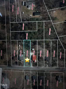 Aerial view of a residential area showing multiple rectangular plots marked with red numbers. A yellow star highlights one plot featuring a red-roofed house. Surrounding areas have trees, additional homes, and roads.
