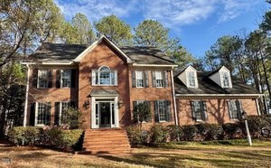 The two-story brick house at 1809 Waddill Way in Henderson boasts multiple windows and a gabled roof, nestled among tall trees. Its central front entrance is highlighted by steps leading up to it. A clear sky and the sun's rays illuminate the welcoming facade of this charming home.